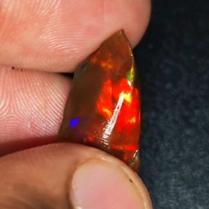 Puede incluir: Una gema de ópalo en bruto con una base de color marrón oscuro. El ópalo muestra un fuego interno vibrante con destellos de rojo, naranja, amarillo, verde y azul. La gema se sostiene entre los dedos, mostrando su forma natural y en bruto.