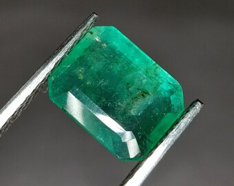 May include: A vibrant green emerald gemstone, cut into a rectangular shape, is displayed on a pair of metal tweezers. The emerald has a slightly cloudy appearance, with some inclusions visible.
