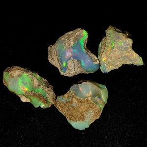Puede incluir: Cuatro piedras de ópalo natural en bruto con colores verde, azul e iridiscente. Las piedras están engastadas en una matriz de roca marrón y gris.