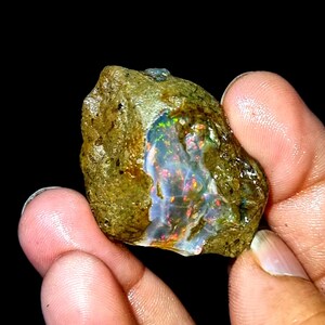 Peut inclure: Une pierre d'opale brute, non taillée, avec un jeu de couleurs vives. La surface de l'opale est composée de tons terreux, avec des éclats de couleurs irisées, notamment du rouge, du bleu et du vert. La pierre est tenue dans une main sur fond noir.