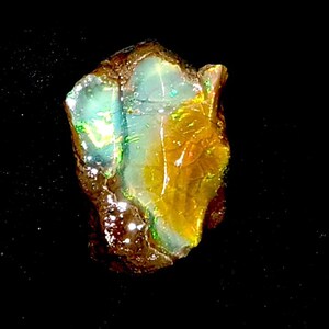 Op de afbeelding: Een ruwe, ongeslepen opaal edelsteen tegen een donkere achtergrond. De opaal toont een mix van kleuren, waaronder geel, groen en blauw, met een iriserend effect. De steen heeft een natuurlijke, onregelmatige vorm.