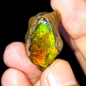Op de afbeelding: Een ruwe, ongeslepen opaal edelsteen met een levendig kleurenspel. De opaal heeft een mix van groene, gele en oranje tinten, met een donkere, aardse matrix. De edelsteen wordt tussen vingers gehouden tegen een zwarte achtergrond.