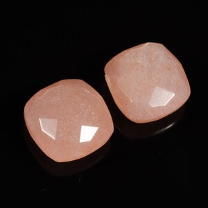 Puede incluir: Dos gemas cuadradas facetadas, de color melocotón y aspecto brillante. Las gemas se muestran sobre un fondo negro, resaltando sus superficies reflectantes. Adecuadas para la creación de joyas.
