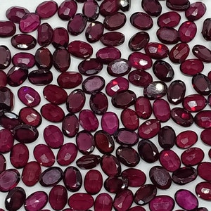 May include: A collection of numerous loose, oval-shaped, faceted gemstones in deep shades of red and burgundy. Each stone features a polished surface with multiple geometric facets that reflect light, creating a uniform appearance across the group.