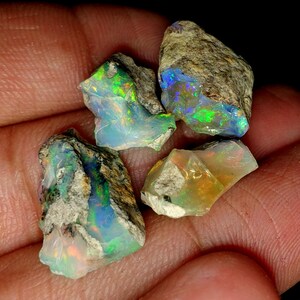 Op de afbeelding: Vier ruwe opaalstenen met iriserende kleuren, waaronder groen, blauw en oranje, in een hand.