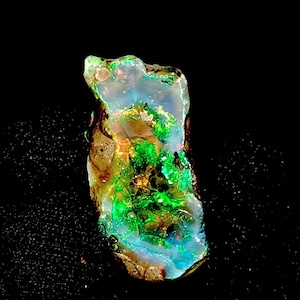 Op de afbeelding: Een levendige, ruwe opaal edelsteen met een opvallende kleurenpracht. De steen heeft een mix van groene, blauwe en gouden tinten, met een doorschijnende kwaliteit. De opaal staat tegen een zwarte achtergrond, wat de natuurlijke schoonheid benadrukt.