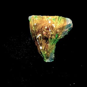 Op de afbeelding: Een abstracte glazen sculptuur in amber met groene en blauwe accenten. Het stuk heeft een textuuroppervlak en een onregelmatige vorm, tegen een zwarte achtergrond. De sculptuur lijkt verlicht te zijn.