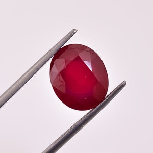 Op de afbeelding: Een gefacetteerde, ovaalvormige robijn edelsteen, vastgehouden door een pincet. De robijn heeft een diepe, rijke rode kleur, met meerdere facetten die het licht vangen. De pincet is zilverkleurig en houdt de edelsteen tegen een witte achtergrond.