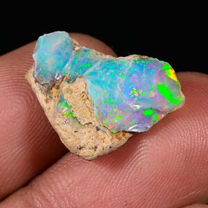 Peut inclure: Une pierre d'opale brute aux couleurs vives. L'opale présente des éclats de vert, de bleu et de jaune sur une base blanc laiteux. La pierre est sertie dans une matrice de roche de couleur beige, mettant en valeur sa forme naturelle.