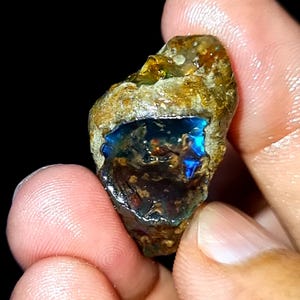 Op de afbeelding: Een ruwe opaal edelsteen met een levendig blauw interieur en een bruine, getextureerde buitenkant. De opaal wordt tussen vingers gehouden tegen een donkere achtergrond, wat de natuurlijke schoonheid en unieke kleurvariaties benadrukt.