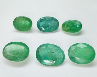 May include: Six oval-shaped green gemstones, likely emeralds, are arranged on a white surface. The gemstones are faceted and have a slight shimmer.