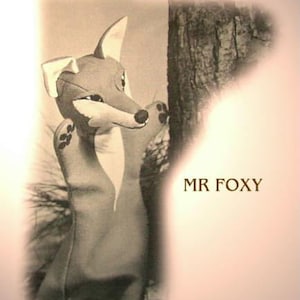 以下が含まれることがあります： 白い口吻と茶色の鼻を持つ灰色のキツネの人形。人形は木の幹の前に立っています。画像の右側に茶色の文字で「MR. FOXY」と書かれています。
