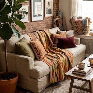 Könnte beinhalten: Ein beiges Sofa mit einer mehrfarbig gestreiften Decke, einem grünen Kissen, zwei gemusterten orangefarbenen Kissen und einem dunkelroten Samtkissen. Ein hölzerner Couchtisch trägt Bücher und eine Tasse, während links eine große Topfpflanze steht.