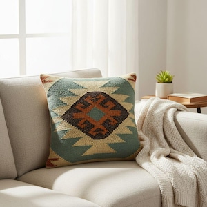 May include: A square throw pillow featuring a textured, woven geometric pattern in shades of teal, cream, and dark brown. The design consists of a central diamond motif surrounded by jagged, stepped shapes that extend toward the pillow edges.