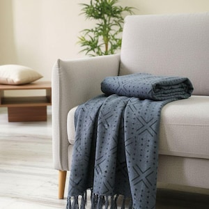 May include: A slate blue throw blanket featuring a dark gray geometric pattern of X shapes and dots. The blanket is folded and draped over a light gray sofa, showcasing a textured fabric finish and decorative fringe along the bottom edge.