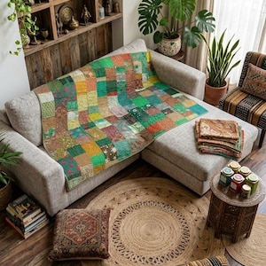 Op de afbeelding: Een patchwork quilt met een raster van veelkleurige stoffen vierkanten in tinten groen, oranje en bruin, gedrapeerd over een lichtgrijze hoekbank. De quilt vertoont gevarieerde texturen en patronen, met een stapel gevouwen stoffen ernaast.