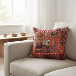 Peut inclure: Un coussin carré présentant un motif patchwork composé de divers panneaux de tissu rouges, orange et marron. La surface est richement ornée de perles complexes, de broderies métalliques et de petits coquillages cauris blancs sur les bords.