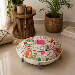 May include: A round floor cushion featuring a patchwork design of various floral and geometric patterns in vibrant colors like pink, green, and red. The cushion has a cream-colored base and is decorated with small, colorful fabric tassels.
