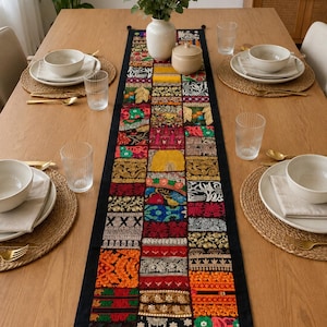 Può includere: Un lungo runner da tavola rettangolare caratterizzato da un design patchwork di vari quadrati di tessuto ricamato in colori vivaci tra cui rosso, arancione, giallo, verde e nero. Il runner presenta bordi neri e piccole nappe nere agli angoli.