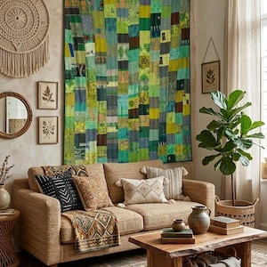 Op de afbeelding: Een grote rechthoekige wandquilt bestaande uit een patchwork van diverse groene, gele en blauwe stoffen vierkanten. De quilt hangt boven een beige bank met diverse kussens met patronen en een geweven deken over de armleuning.