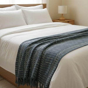 Op de afbeelding: Een donkerblauwe plaid met een wit chevron- en stippenpatroon, gedrapeerd over een bed. De deken heeft een geweven textuur en franjes aan de onderkant, wat contrasteert met het effen witte beddengoed en de kussens.