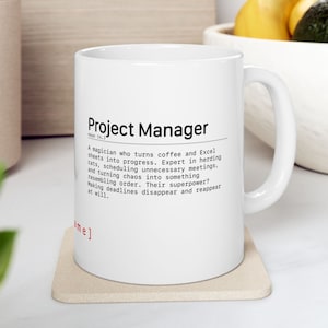 Könnte beinhalten: Weiße Keramik-Tasse mit der Aufschrift "Project Manager" und einer Definition. Die Definition beschreibt einen Projektmanager als einen Zauberer, der Chaos in Ordnung verwandelt. Die Tasse steht auf einem beigen Untersetzer.