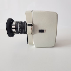 May include: A white vintage 8mm film camera with a black lens. The lens has a zoom ring and a depth of field scale. The camera has a built-in viewfinder.