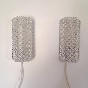May include: Two clear, rectangular wall sconces with a diamond-patterned texture. Each sconce has a ridged top and bottom, and a wire extends from the base. The sconces are mounted on a white wall.