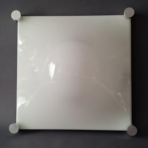 Könnte beinhalten: Quadratische, weiße, glänzende Wandkunst mit vier silbernen, zylindrischen Eckhalterungen. Das Kunstwerk hat eine reflektierende Oberfläche, und die Ecken sind mit kleinen, kreisförmigen Metallbefestigungen gesichert. Das Stück ist auf einem dunkelgrauen Hintergrund montiert.