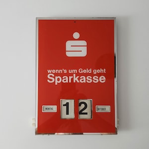 May include: A rectangular red wall calendar with a chrome frame featuring the white Sparkasse logo and the text "wenn's um Geld geht Sparkasse". It displays the date "12" in large black numerals, with "MONTAG" and "OKTOBER" in small windows.