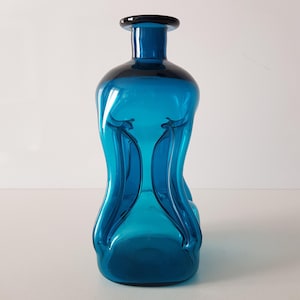 May include: A teal blue glass vase with a unique, organic shape. The vase has a wide base and a narrow neck, with a series of curves and dips in the glass.
