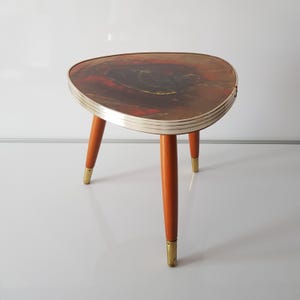 Puede incluir: Una mesa auxiliar de estilo moderno de mediados de siglo con una tapa triangular y tres patas de madera. La tapa está hecha de laminado marrón y naranja con un borde plateado. Las patas tienen puntas de latón.