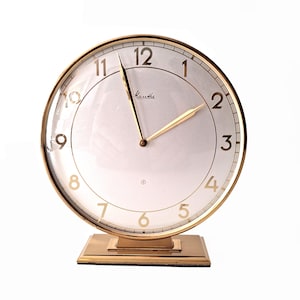 May include: A gold-toned analogue clock with a white face and black Roman numeral hour markers. The clock has a gold-toned base and the brand name "Kauthe" is printed on the face.