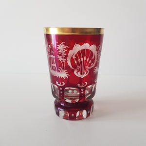 Puede incluir: Un gobelet de vidrio rojo con un borde dorado y diseños florales grabados en blanco. El gobelet tiene una base de vidrio tallado.