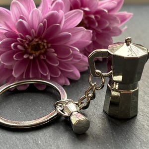 May include: A silver espresso maker keyring with a miniature espresso maker charm. The keyring is attached to a silver ring. Two pink flowers are in the background.