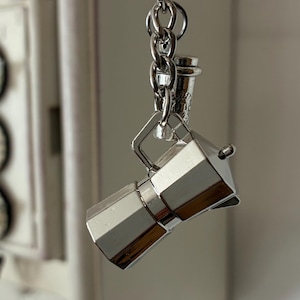 May include: A silver-coloured keychain featuring a miniature espresso maker and a small coffee cup charm. The espresso maker has a classic octagonal design. The keychain is attached to a silver ring and chain.