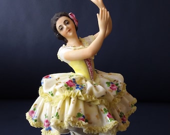 May include: A porcelain figurine of a dancer with dark hair and a pink flower accessory. The dancer wears a yellow bodice and a voluminous, ruffled white skirt decorated with small pink, blue, and green floral patterns and yellow trim.