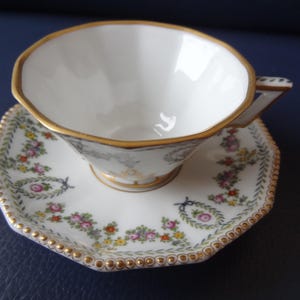Könnte beinhalten: Ein weißes Porzellan-Tassenset mit Untertasse in achteckiger Form und goldfarbenen Rändern. Die Außenseite der Tasse und die Oberfläche der Untertasse sind mit kleinen, bunten Blumengirlanden und Kränzen in Rosa, Gelb und Grün verziert.