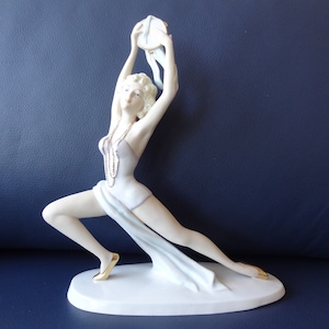 Puede incluir: Una figura de porcelana de una persona bailando en una pose dinámica, con un leotardo blanco con un acento de tela drapeada, zapatos dorados y un collar largo de cuentas. La figura sostiene un objeto circular sobre la cabeza con los brazos.