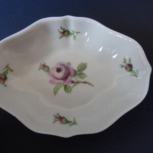 Puede incluir: Un plato ovalado de porcelana blanca con borde festoneado, que presenta una ilustración central de una rosa rosa con hojas verdes y cuatro motivos de capullos de rosa más pequeños colocados simétricamente en el borde interior.