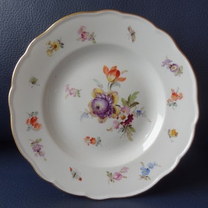 Puede incluir: Plato de porcelana blanca con borde festoneado y ribete dorado. El plato está decorado con un ramo floral central en tonos morados, naranjas y amarillos, y diseños florales más pequeños. También se ve una mariposa.