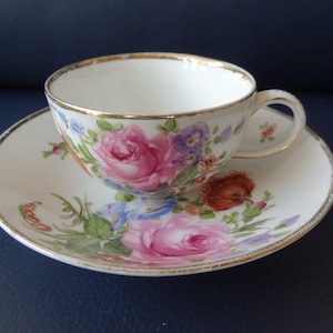 May include: A white teacup and saucer set with a floral design featuring pink roses, purple pansies, and blue accents. The rims are trimmed with gold. The teacup has a handle and sits on a matching saucer.