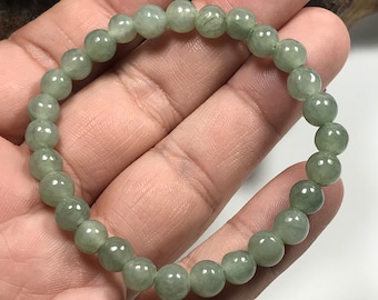 May include: A light green beaded bracelet. The bracelet is made of round, polished beads. The beads have a translucent appearance and a subtle, natural variation in colour. The bracelet is held in a hand.