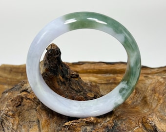 May include: A jade bangle bracelet with a smooth, polished surface. The bracelet features a blend of white and green hues, creating a marbled effect. The jade is displayed on a piece of textured, brown wood.