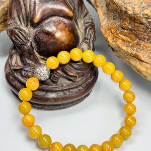 Pode incluir: Uma pulseira de contas redondas amarelo-alaranjadas. A pulseira tem um fecho prateado. As contas s&atilde;o enfiadas para formar um c&iacute;rculo completo. A pulseira &eacute; exibida em frente a uma est&aacute;tua de madeira.