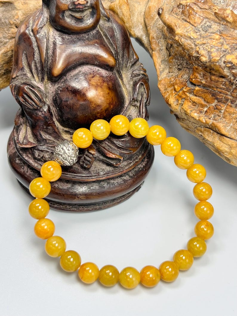 Pode incluir: Uma pulseira de contas redondas amarelo-alaranjadas. A pulseira tem um fecho prateado. As contas s&atilde;o enfiadas para formar um c&iacute;rculo completo. A pulseira &eacute; exibida em frente a uma est&aacute;tua de madeira.