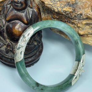 May include: A jade bangle bracelet with silver accents. The bracelet is a light green color with darker green and black inclusions. The silver accents feature an ornate design. The bracelet is displayed in front of a brown statue and a piece of wood.