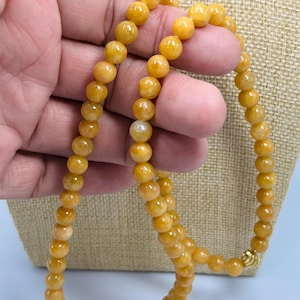 Könnte beinhalten: Eine Halskette aus goldgelben Perlen. Die Perlen sind rund und scheinen aus poliertem Stein zu bestehen. Die Halskette wird von einer Hand gehalten, und der Verschluss ist goldfarben. Der Hintergrund ist neutral beige.