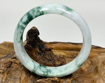 May include: A jade bangle bracelet with a marbled appearance. The bracelet features a mix of white and green hues, with the green color forming a swirling pattern throughout the stone. The bracelet is displayed on a piece of wood.