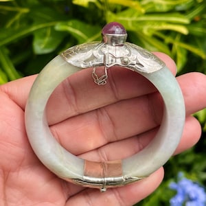 May include: A jade bangle bracelet with a silver clasp and a small, round, red gemstone on top. The jade is a pale green color. The bracelet is held in a hand against a background of green foliage.